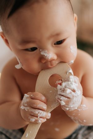 Messy baby enjoying a cake smash, holding a wooden spoon covered in white frosting with cake on face and hands.