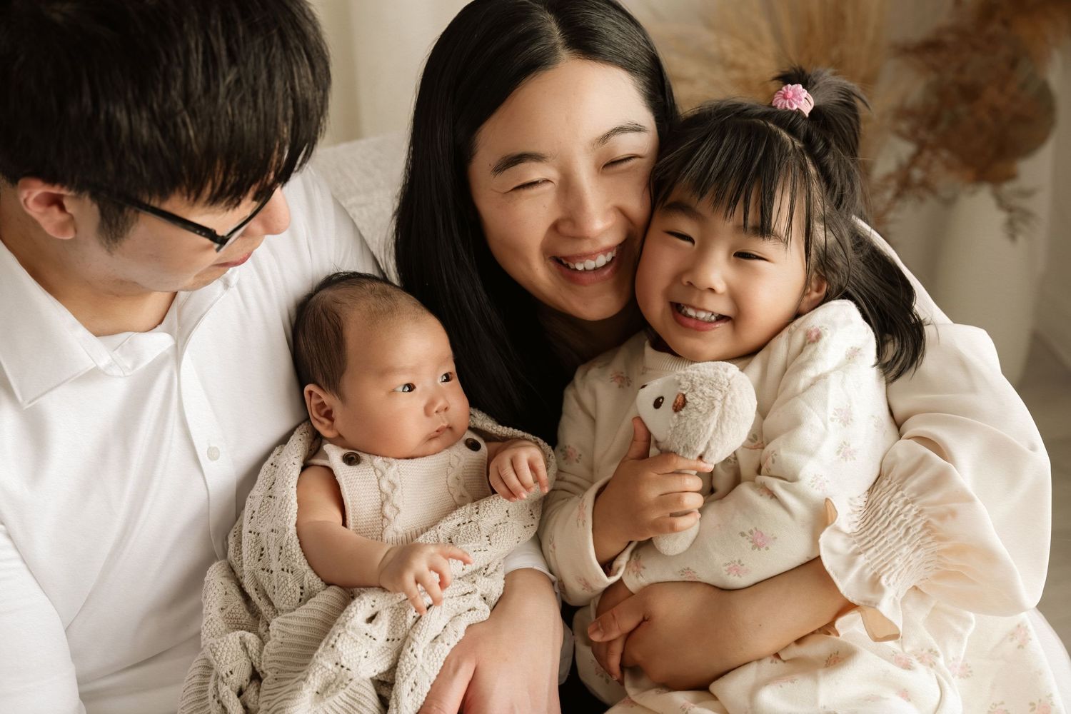 Happy Asian family portrait with parents holding newborn baby and smiling toddler girl in cozy white outfits.
