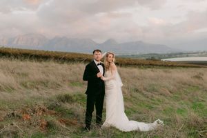 Bride and groom pose in a scenic field with misty mountains at sunset, bride in white gown, groom in black tuxedo.