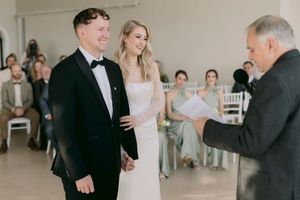 Bride and groom holding hands during wedding ceremony while officiant reads vows in elegant white venue.