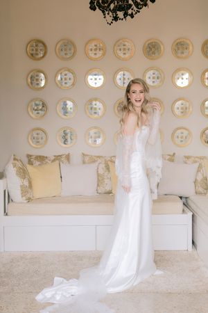Smiling bride in elegant white gown with lace wrap stands in a luxurious room with gold circular mirror wall decor.