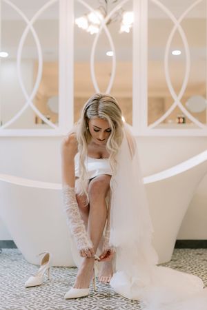 Bride in white wedding dress and veil adjusting her heels while seated near a freestanding bathtub.
