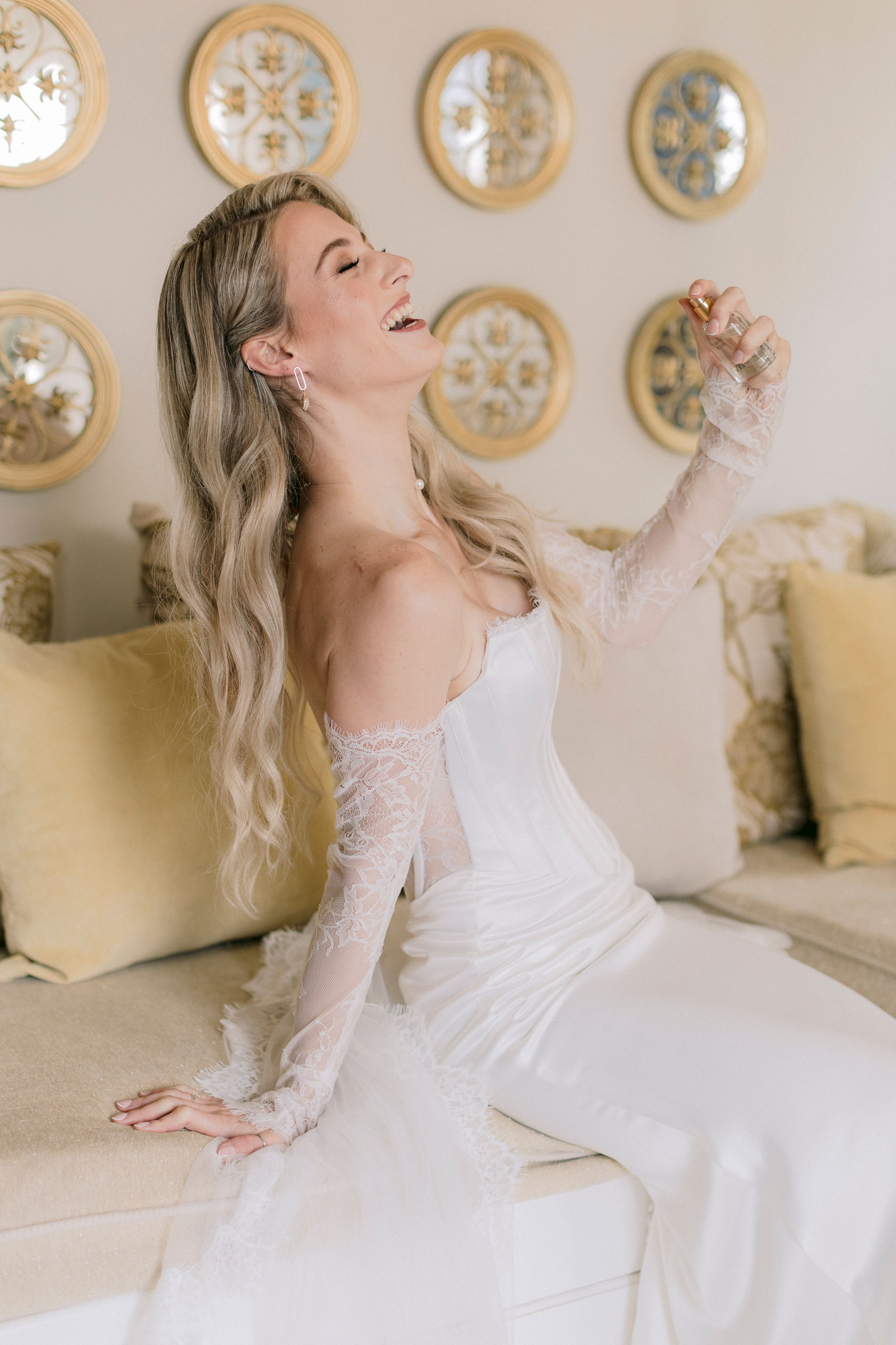 Bride in white lace wedding gown sitting on elegant sofa, laughing joyfully with long blonde waves and drop earrings.