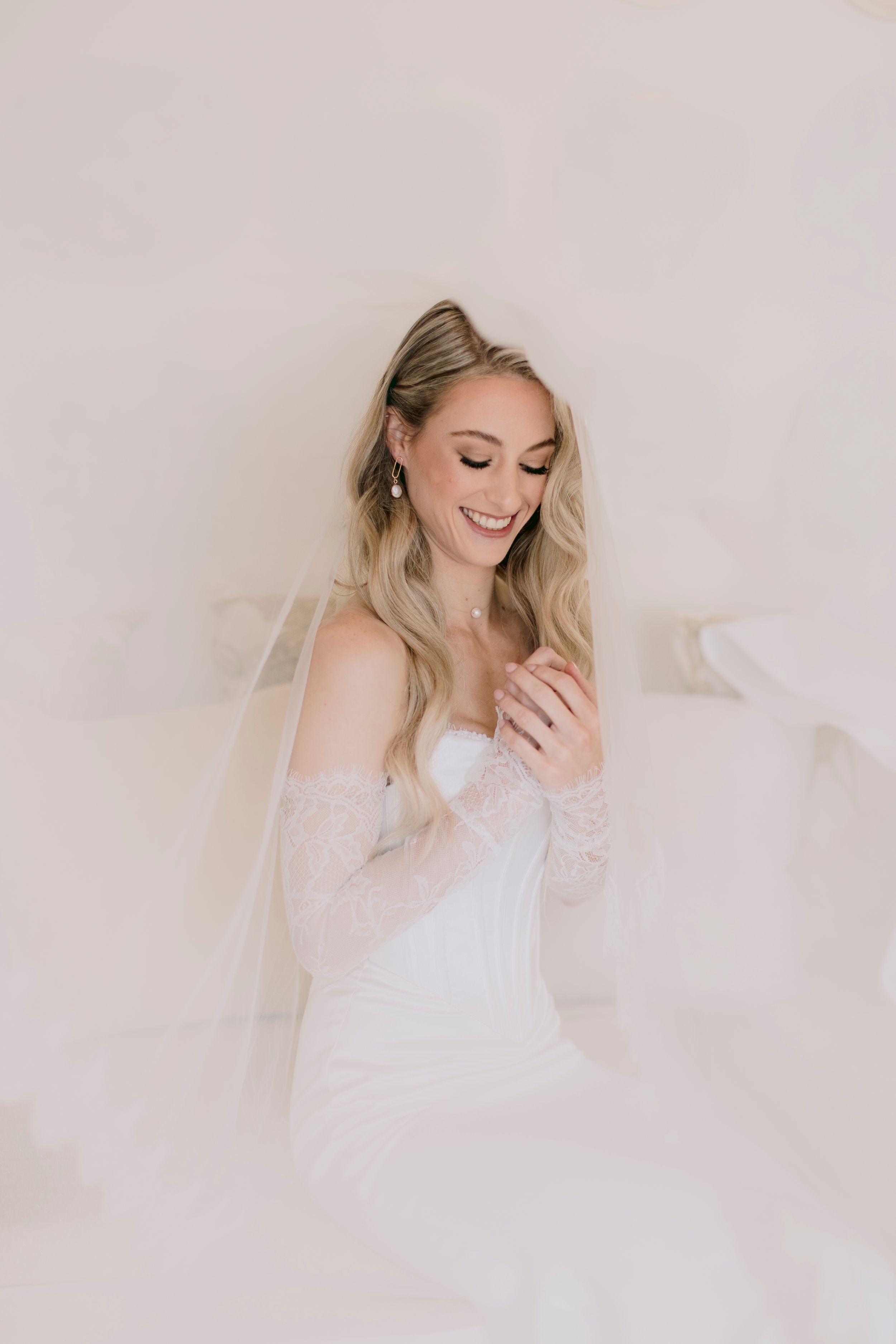 Smiling bride in white strapless gown with lace sleeves and veil, looking down gracefully in soft light.