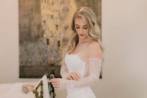 Elegant bride in off-shoulder lace wedding gown adjusting her sleeve, with golden artwork in background.