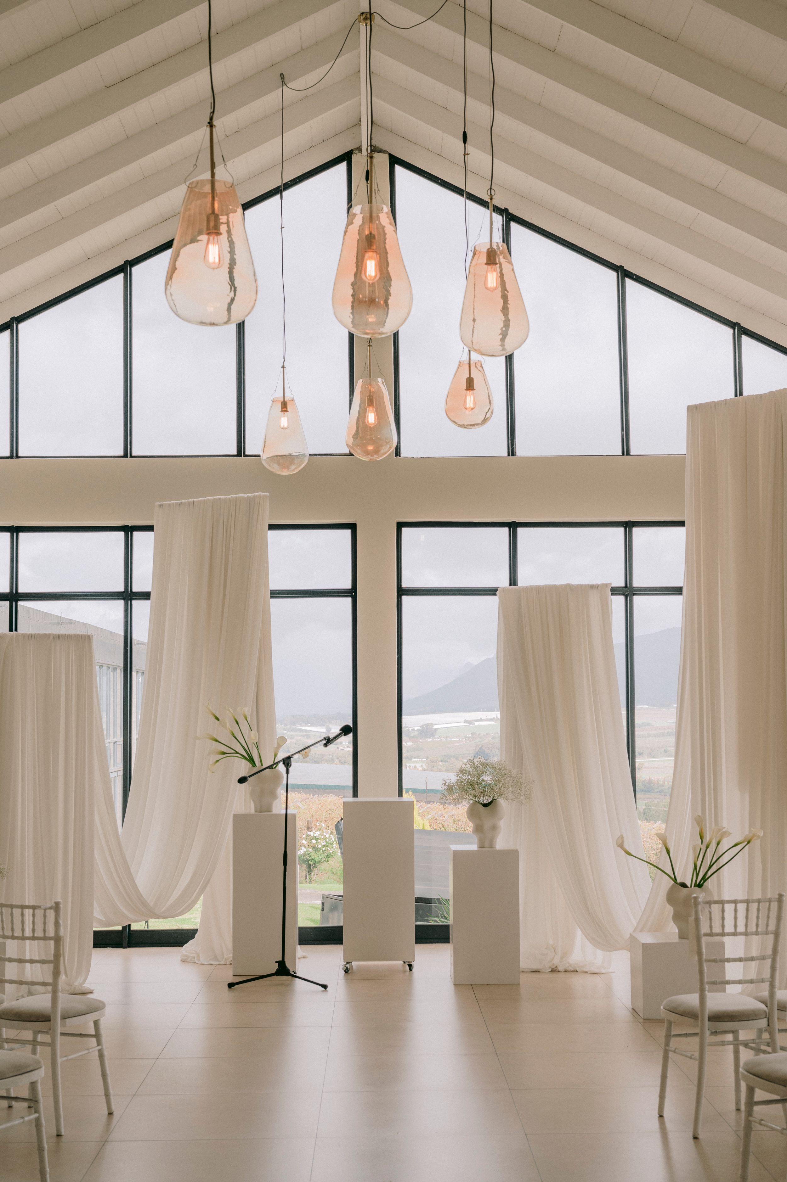 Elegant wedding ceremony venue with white draping, pendant lights, and floor-to-ceiling windows overlooking scenic mountains.