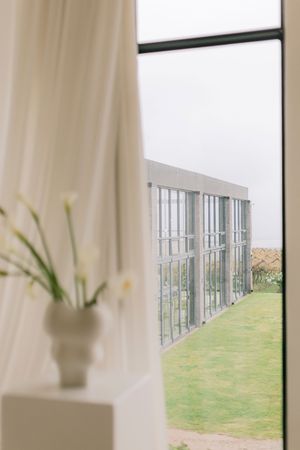 White vase with flowers on windowsill overlooking a greenhouse and green lawn through sheer curtains.