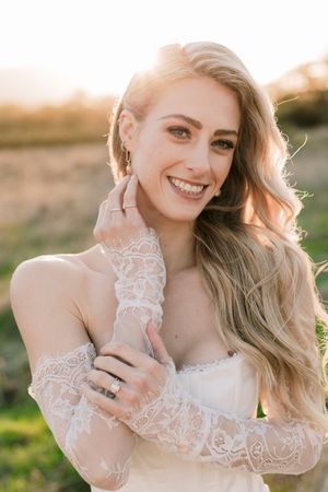 Smiling bride in lace gloves and gown poses outdoors at golden hour sunset with long wavy blonde hair.