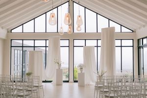 Elegant white wedding ceremony venue with draped fabric columns, ghost chairs, and pendant lights beneath a vaulted ceiling.