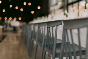 Gray wooden chairs lined up along a long white table in a restaurant with warm bokeh string lights.