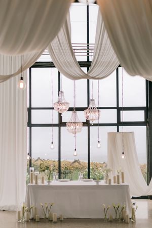 Elegant wedding reception sweetheart table with white draping, crystal chandeliers, candles, and large windows overlooking greenery.