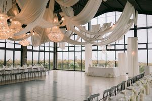 Elegant wedding reception hall with white draped ceiling, crystal chandeliers, long banquet tables, and floor-to-ceiling windows.