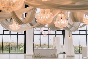 Elegant wedding reception hall with crystal chandeliers, white draped ceiling, and a decorated sweetheart table by large windows.
