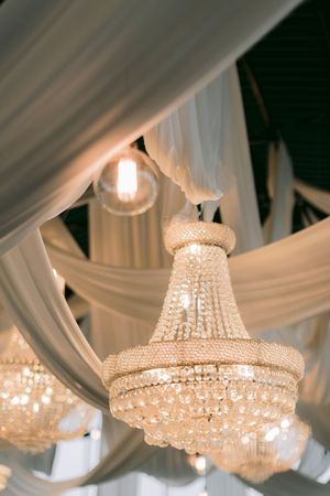 Elegant crystal chandelier hanging from draped white fabric ceiling at a luxury wedding venue.
