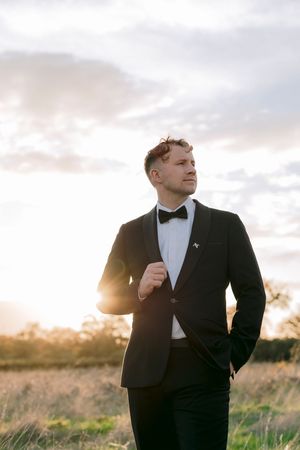 Man in black tuxedo with bow tie standing in sunlit field at golden hour, looking confidently into the distance.