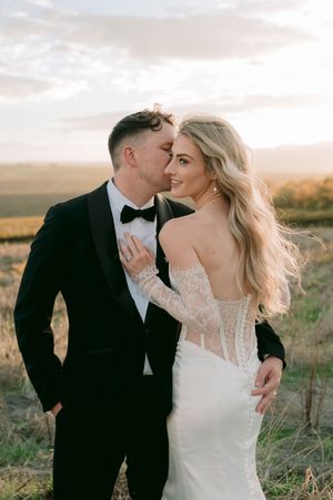A groom in a black tuxedo kisses his blonde bride in a lace backless wedding gown at golden hour outdoors.