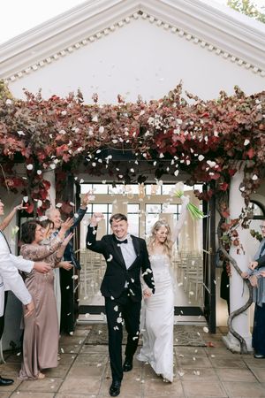 Bride and groom exit wedding ceremony under floral arch as guests toss white flower petals in celebration.
