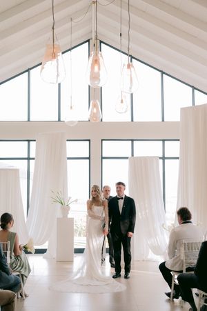 Bride and groom stand at elegant indoor wedding ceremony with white drapes and hanging glass pendant lights.