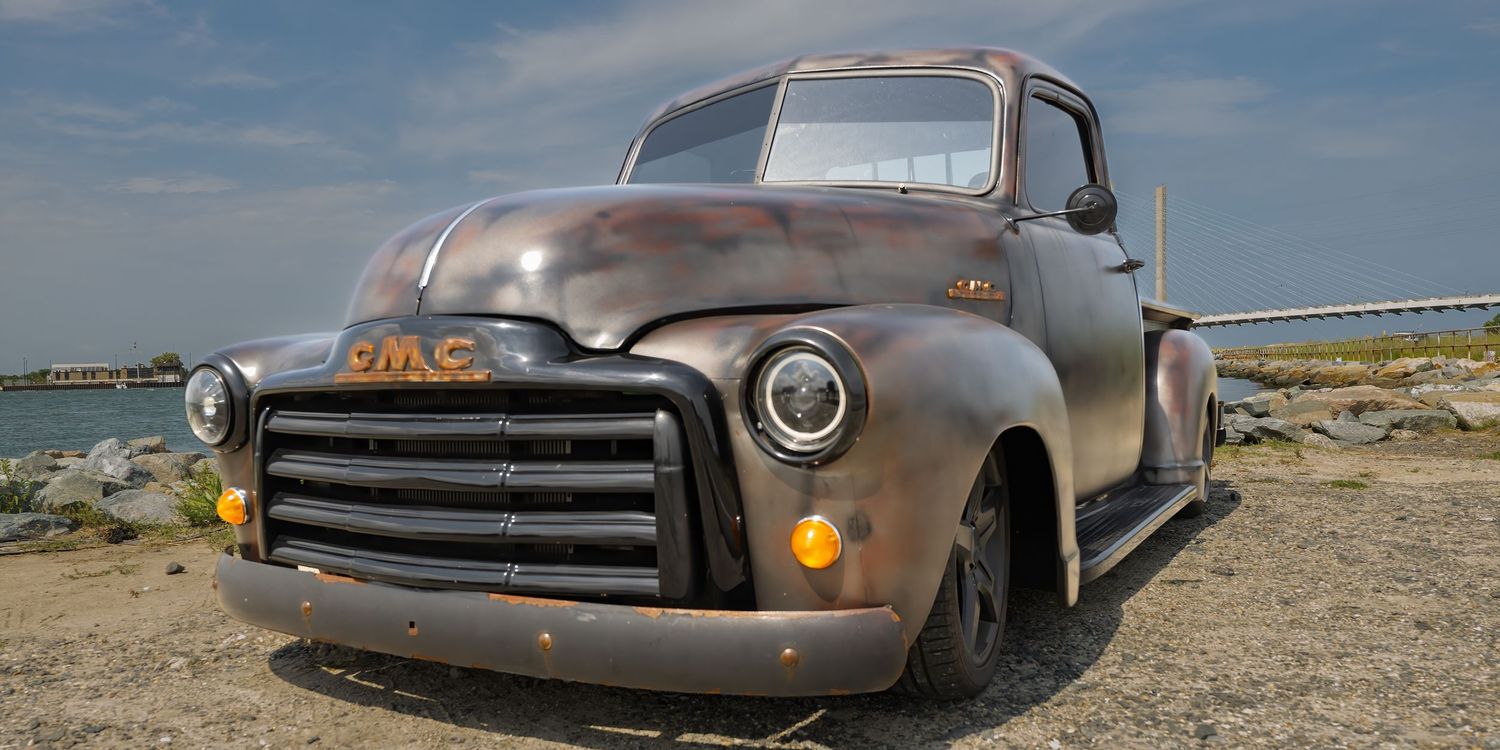 Vintage rusty GMC pickup truck parked on rocky shoreline with ocean and bridge in background.