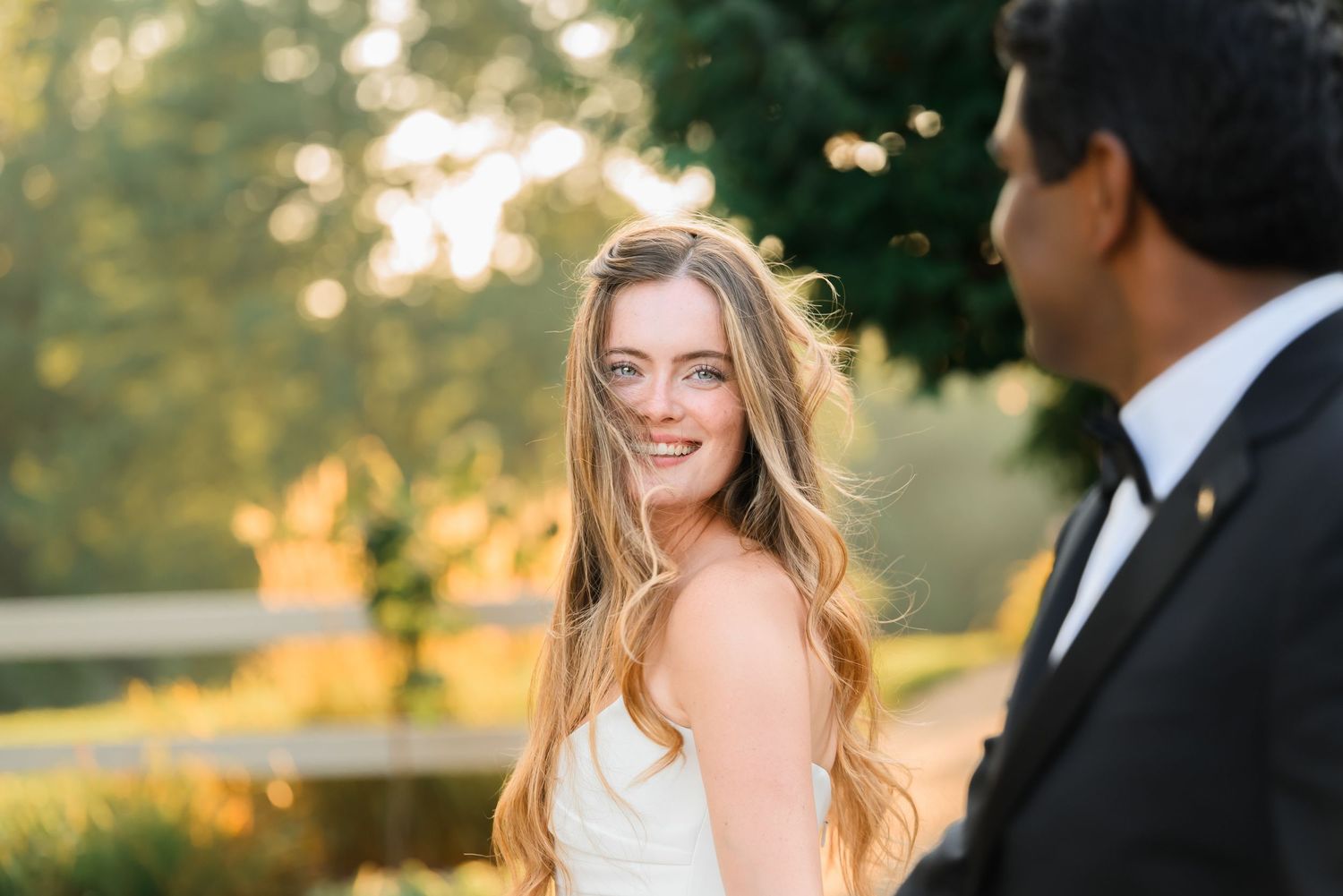Bride smiling over her shoulder in golden hour light, wearing a white gown with flowing hair at outdoor wedding.