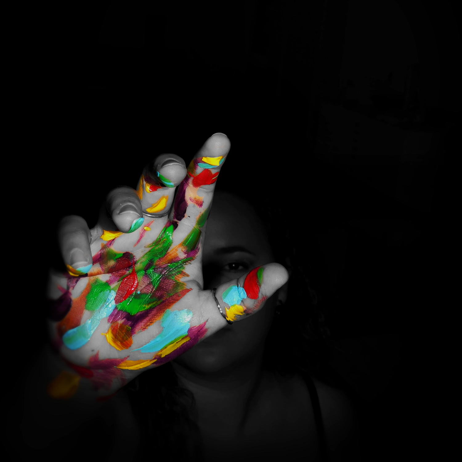 A woman holds up a paint-covered hand toward the camera in a dramatic black and white photo with colorful splashes.