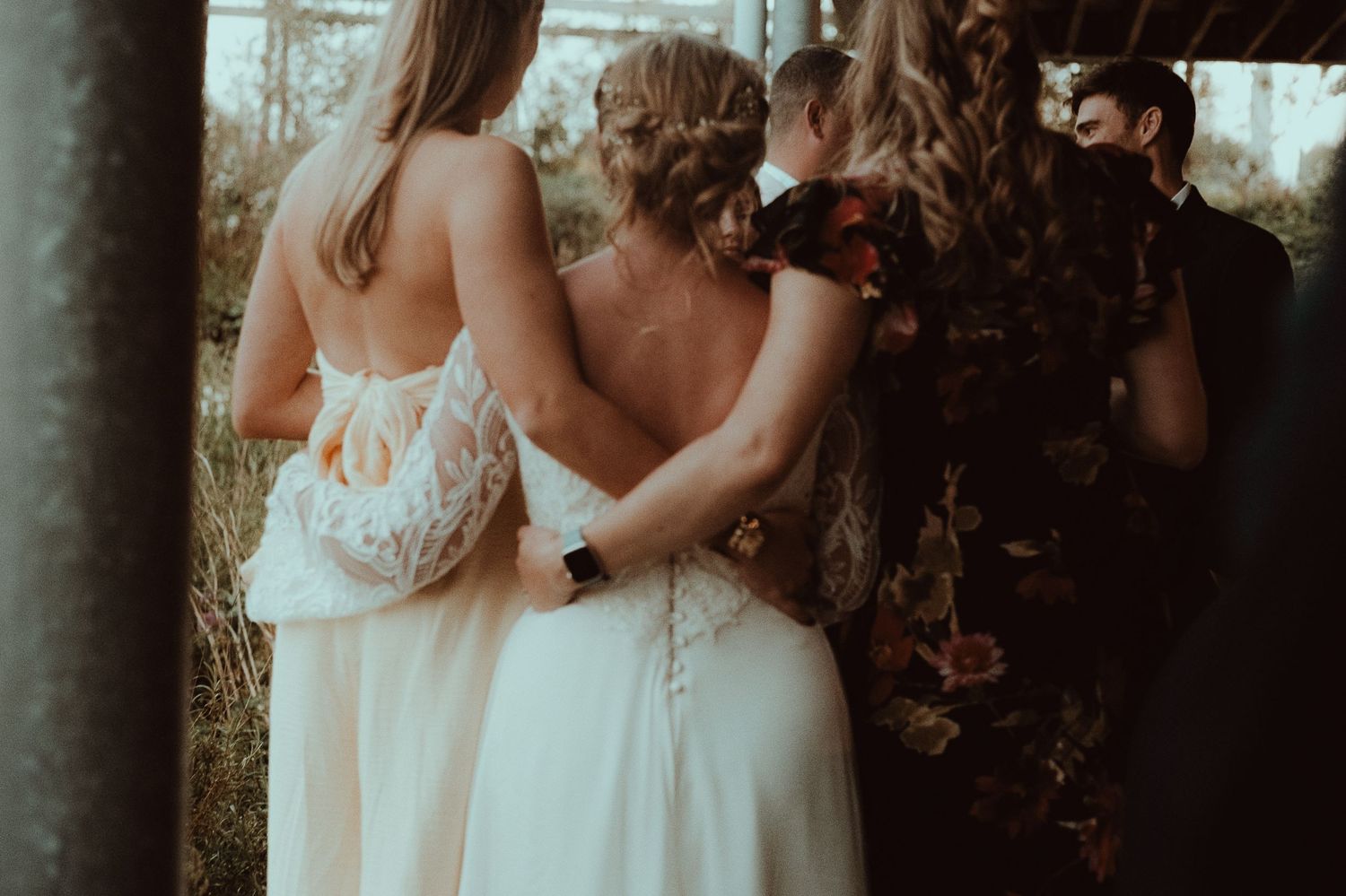 Bride in white lace gown embraced by bridesmaid during outdoor wedding ceremony with floral arch.