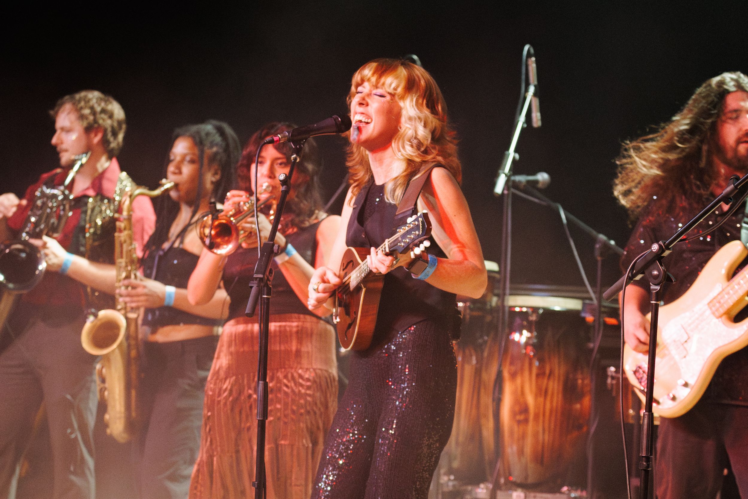 Live band performance with female guitarist, saxophonist, trumpet player, and guitarist on stage under bright lights.