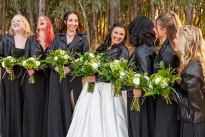 Bride in white gown with bridesmaids in black dresses and leather jackets holding white and green bouquets outdoors.