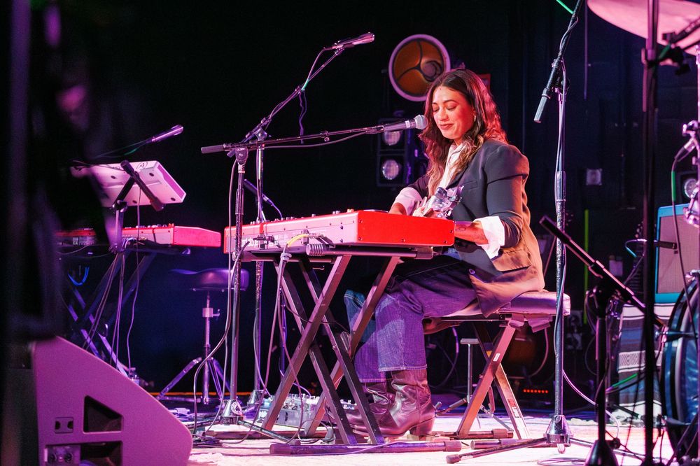 Woman playing red keyboard synthesizer on stage during live music performance under colorful stage lighting.