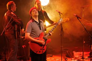 Band performing live on stage, guitarist in red hat playing electric guitar under dramatic orange stage lighting.