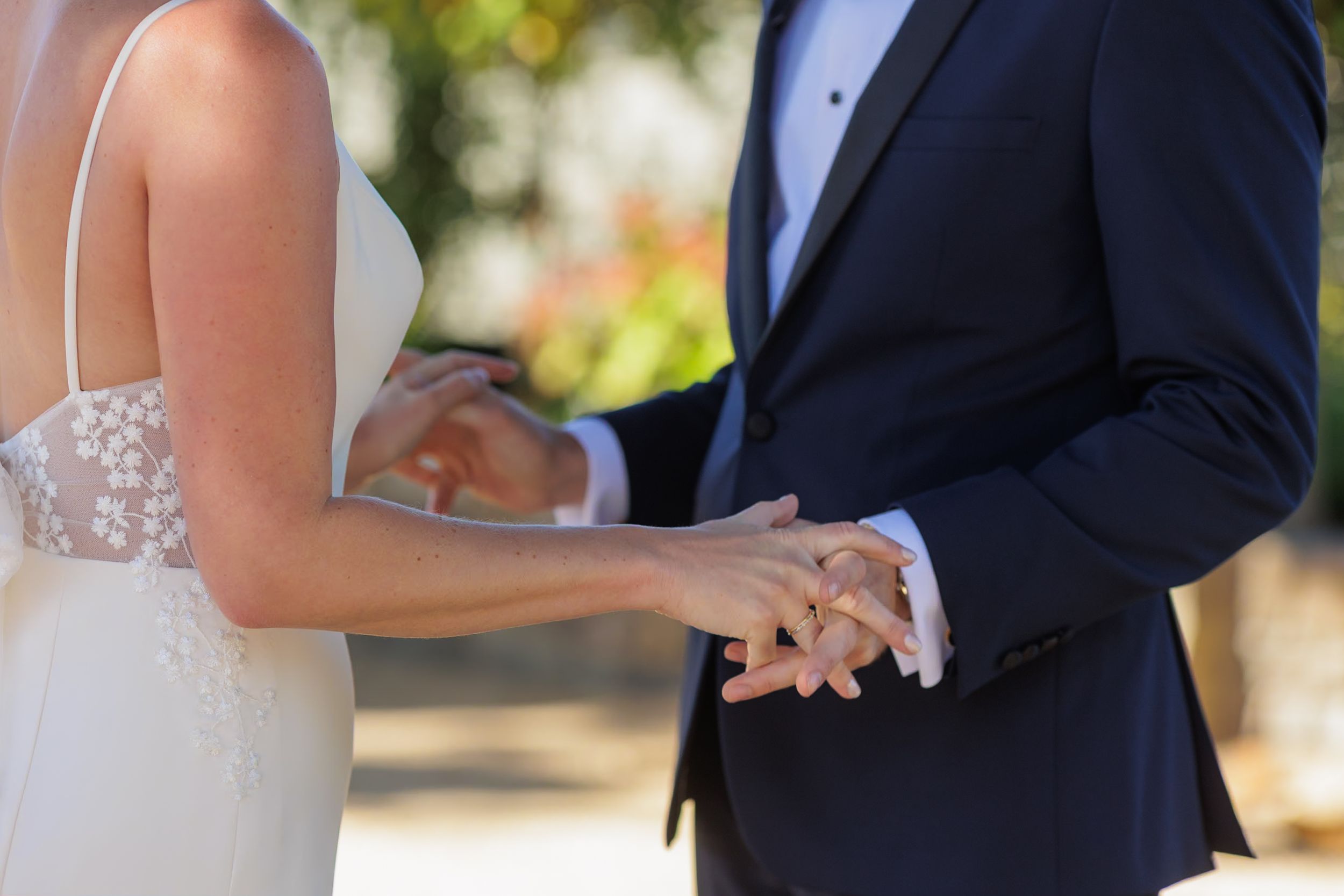 Bride and groom exchanging rings during outdoor wedding ceremony, bride in lace gown, groom in navy suit.
