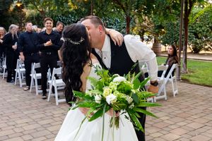 Bride and groom share a kiss at outdoor wedding ceremony with white chairs and guests in background.