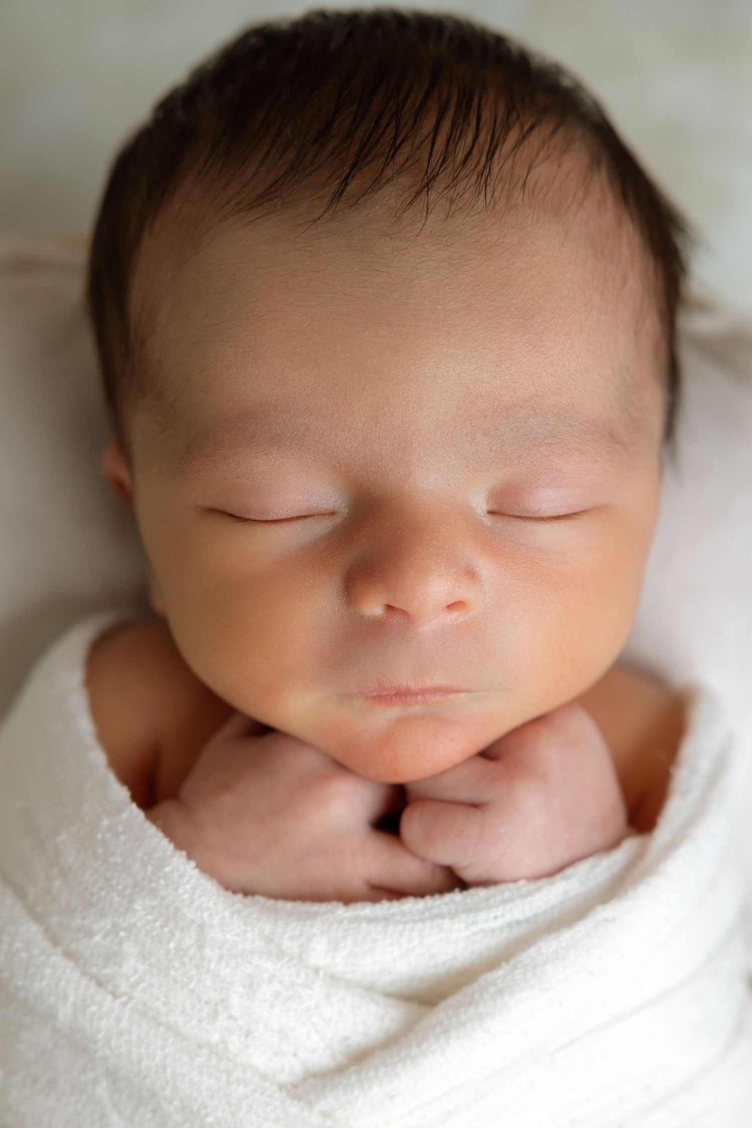 Peaceful newborn baby swaddled in white wrap sleeping with tiny hands tucked under chin.