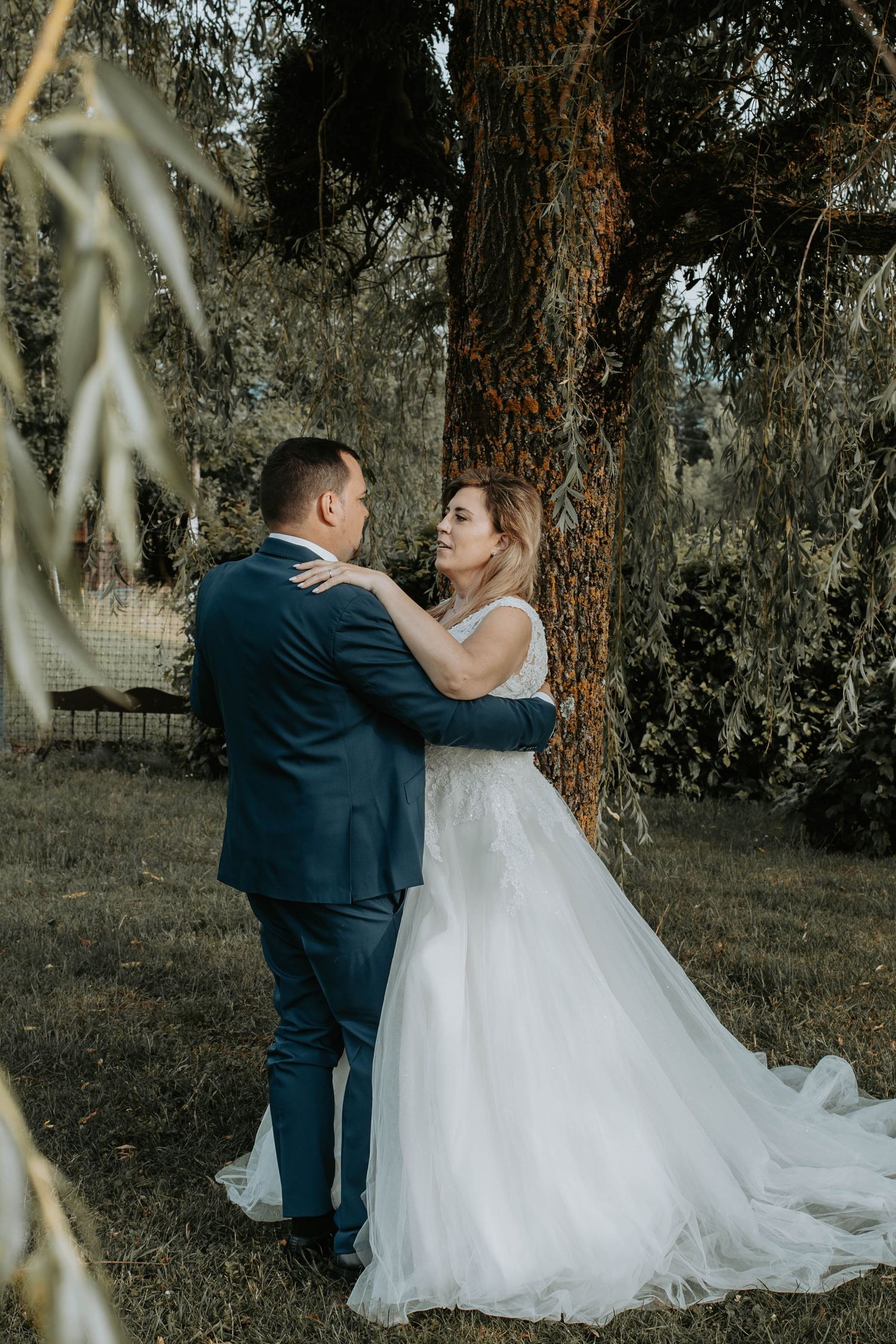 A bride in a white tulle gown and groom in a navy suit embrace romantically beneath a large tree outdoors.