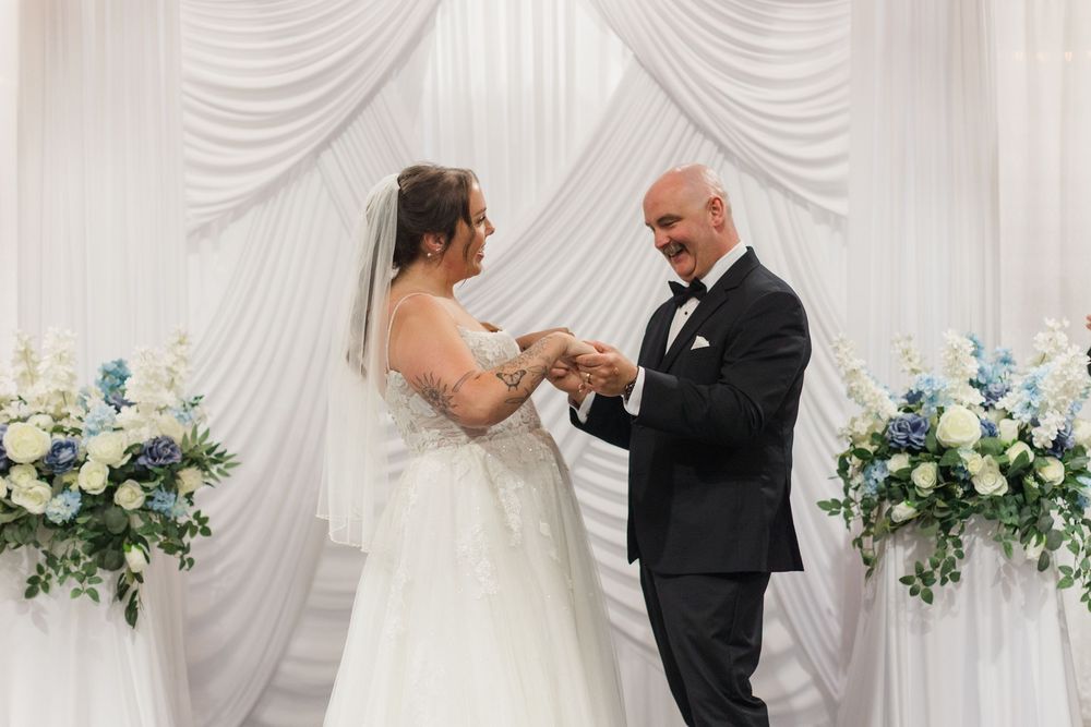 Bride and groom exchange rings at elegant wedding ceremony with white drape backdrop and blue floral arrangements.