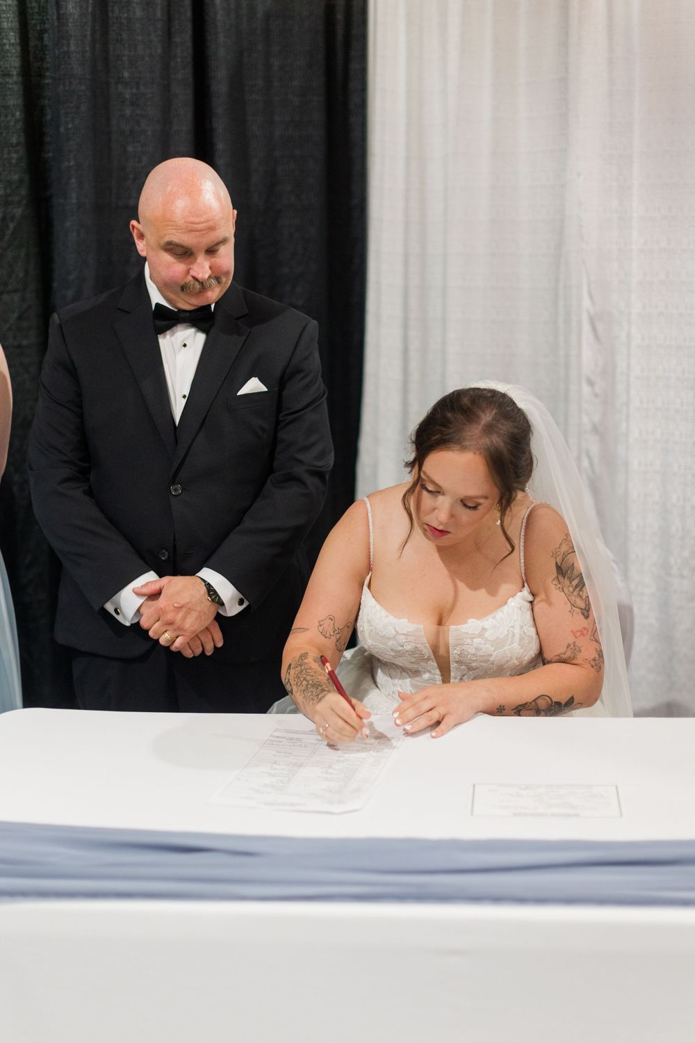 Bride in lace gown signs wedding certificate as groom in black tuxedo watches during wedding ceremony.