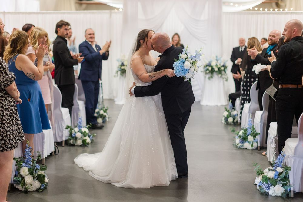 Bride and groom share first kiss at indoor wedding ceremony with guests applauding, blue floral decorations lining the aisle.