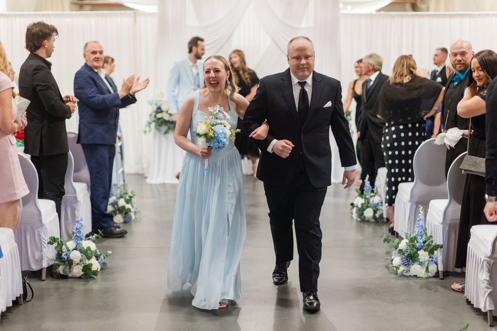 Bride in light blue gown walks down wedding aisle with escort, guests applauding in decorated venue.