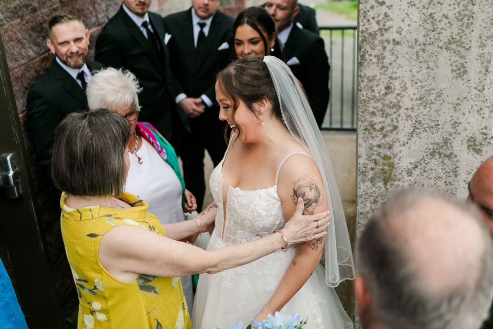 Bride in white lace gown and veil smiles as guests congratulate her outside wedding venue.
