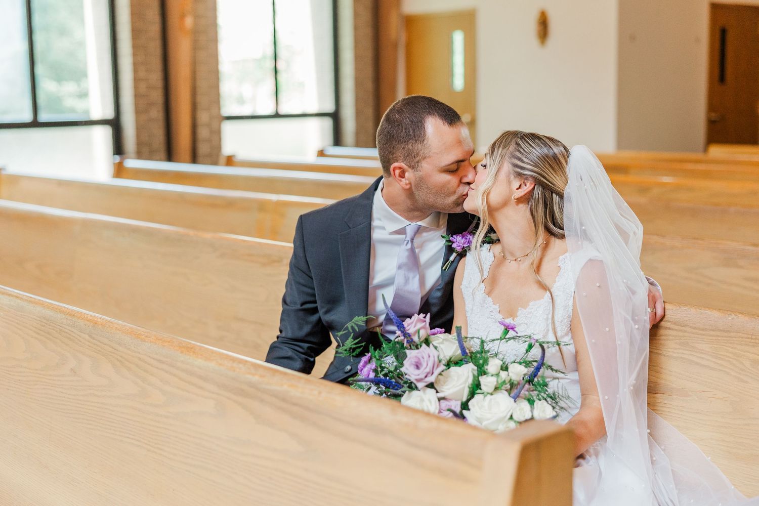 Bride and groom sharing a kiss in wooden church pews, bride holding purple and white bouquet wearing veil.