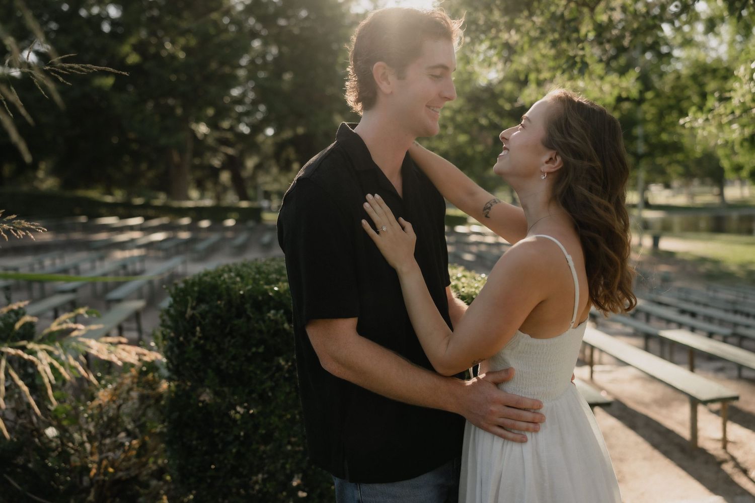 Couple embracing outdoors in golden sunlight, woman in white dress smiling up at man in black shirt near park benches.