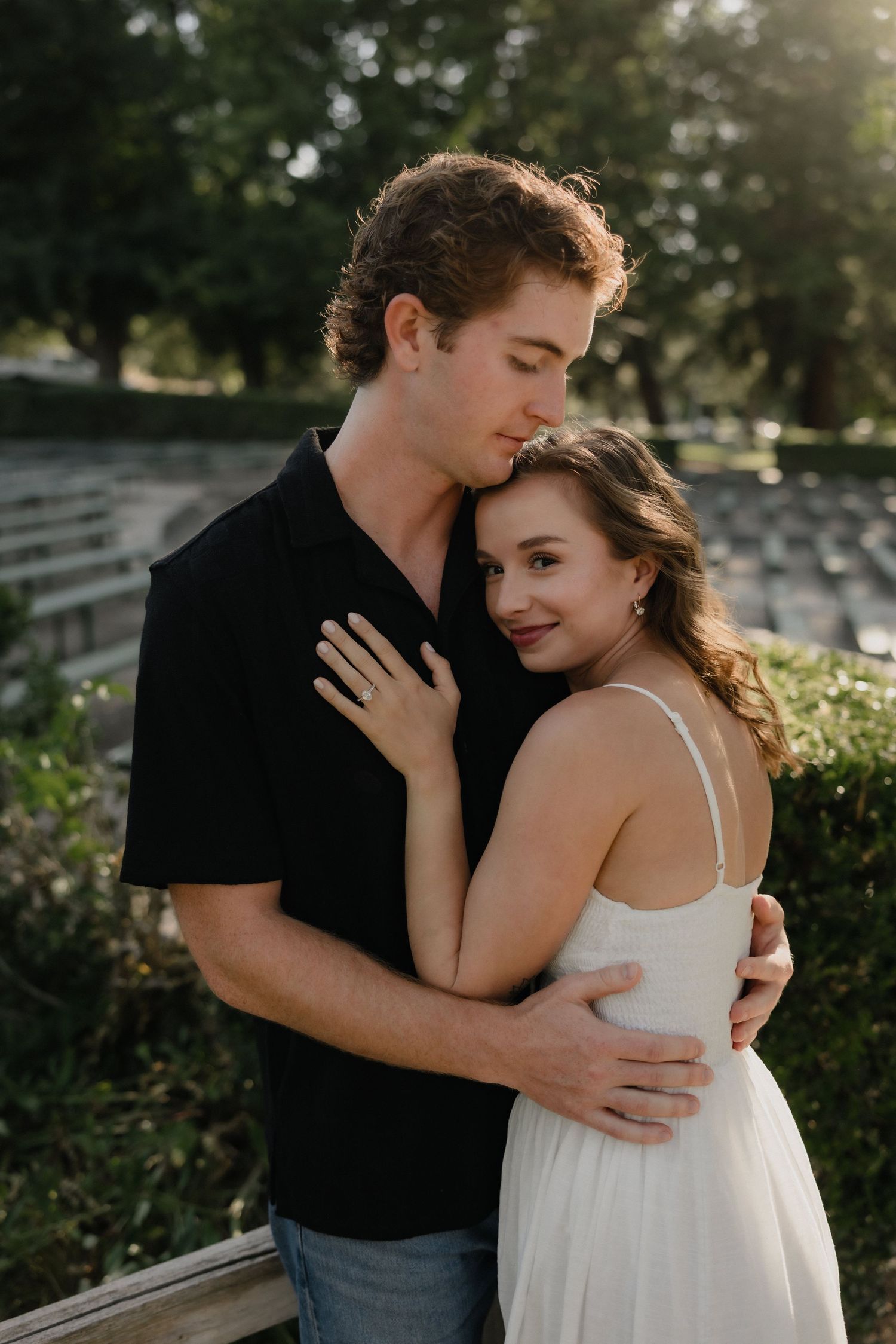 Young couple embracing outdoors at golden hour, woman in white dress smiling, man in black shirt holding her close.