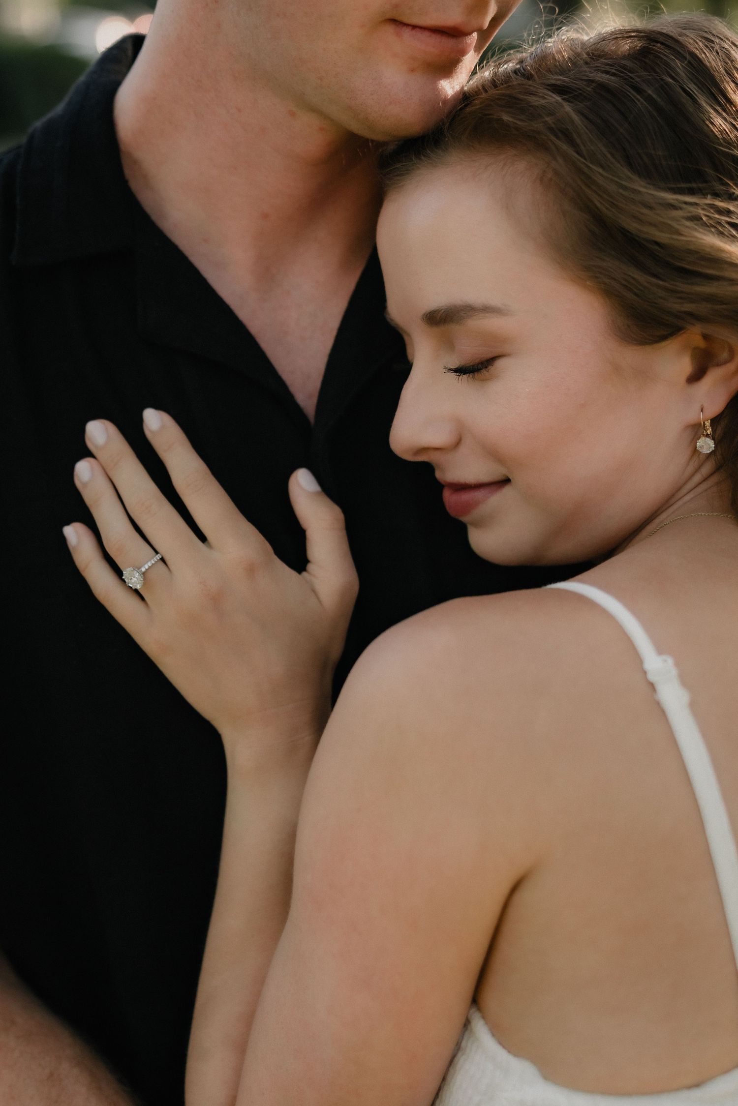 A couple embraces tenderly, the woman wearing an engagement ring and white dress, leaning against a man in a black shirt.