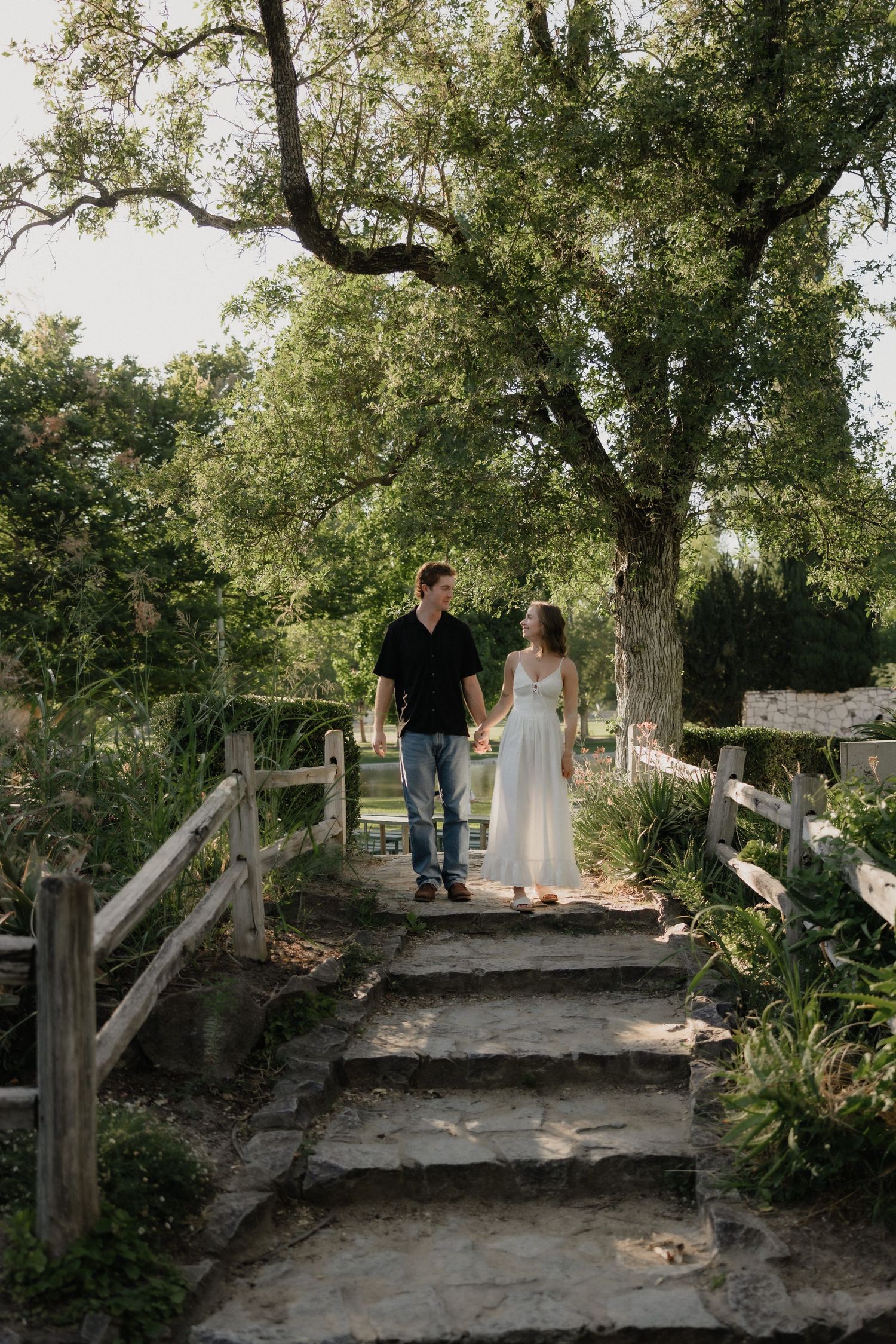 Couple holding hands walking up stone steps surrounded by lush greenery, woman in white dress and man in black shirt.