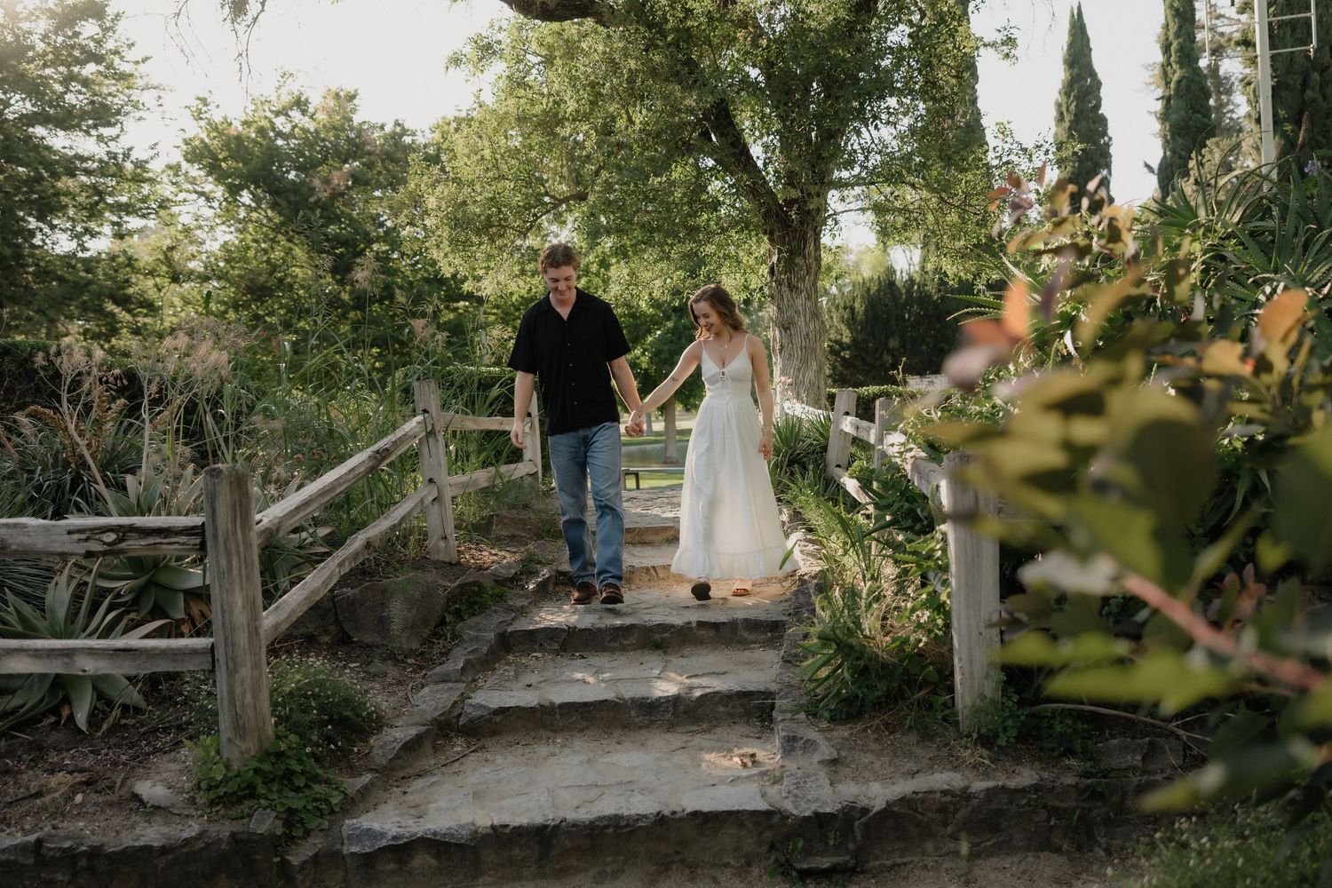 Couple holding hands walking down stone garden steps surrounded by lush greenery, man in black shirt and woman in white dress.