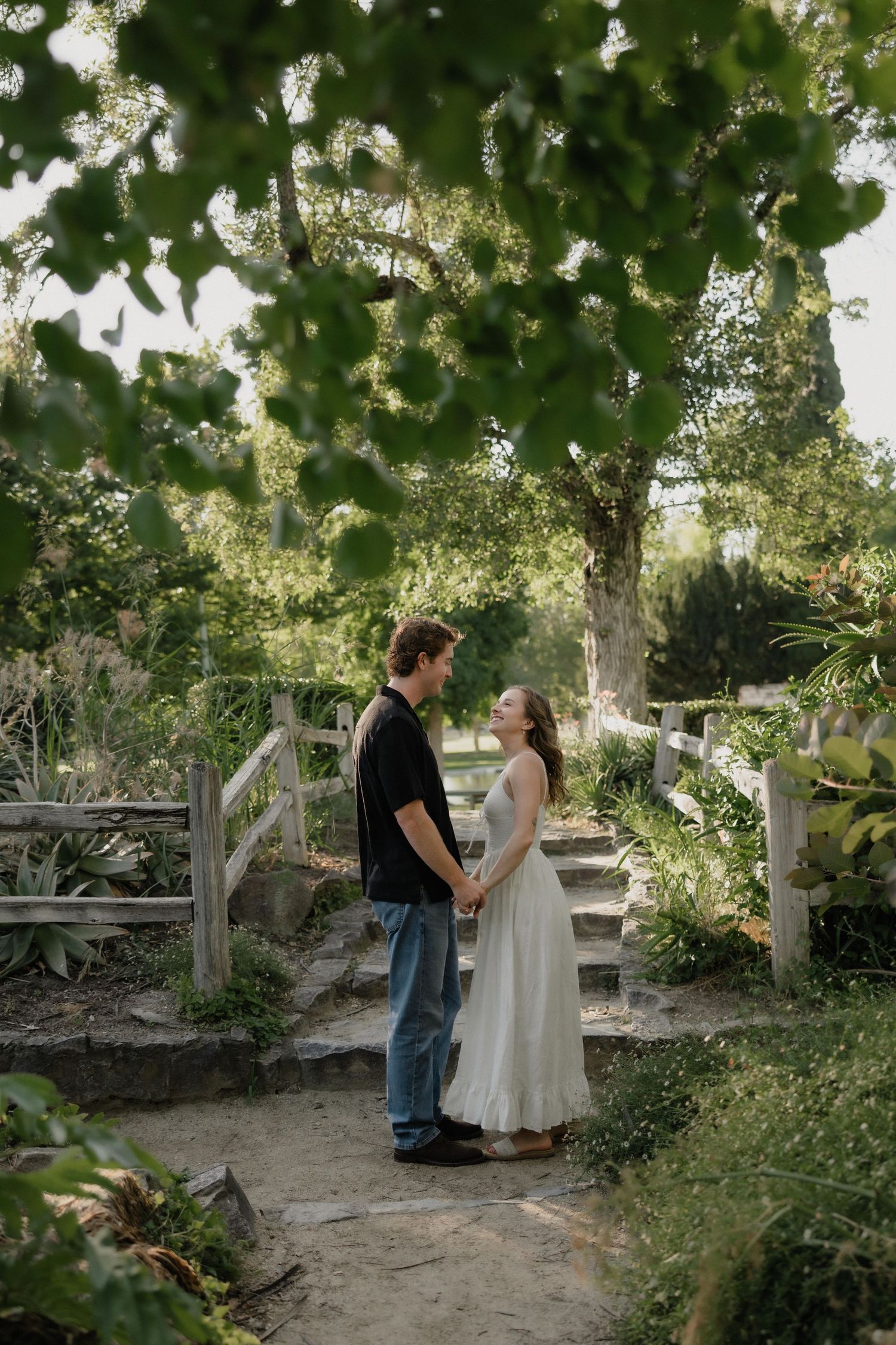 A couple holds hands and gazes at each other on a stone path in a lush garden, framed by green leaves.