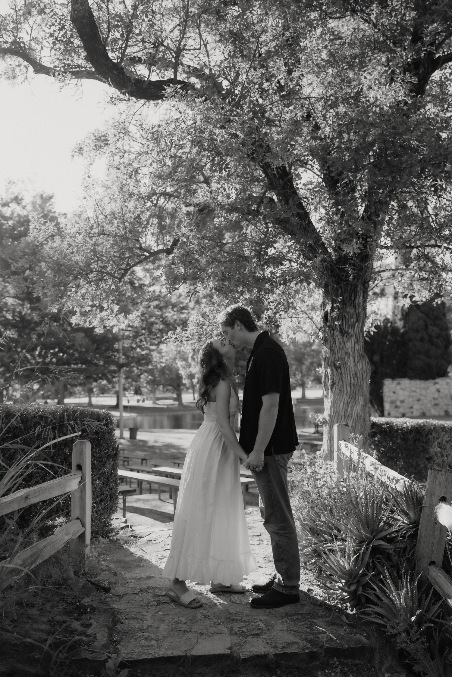 Black and white photo of a couple sharing a romantic kiss in a lush garden park setting.