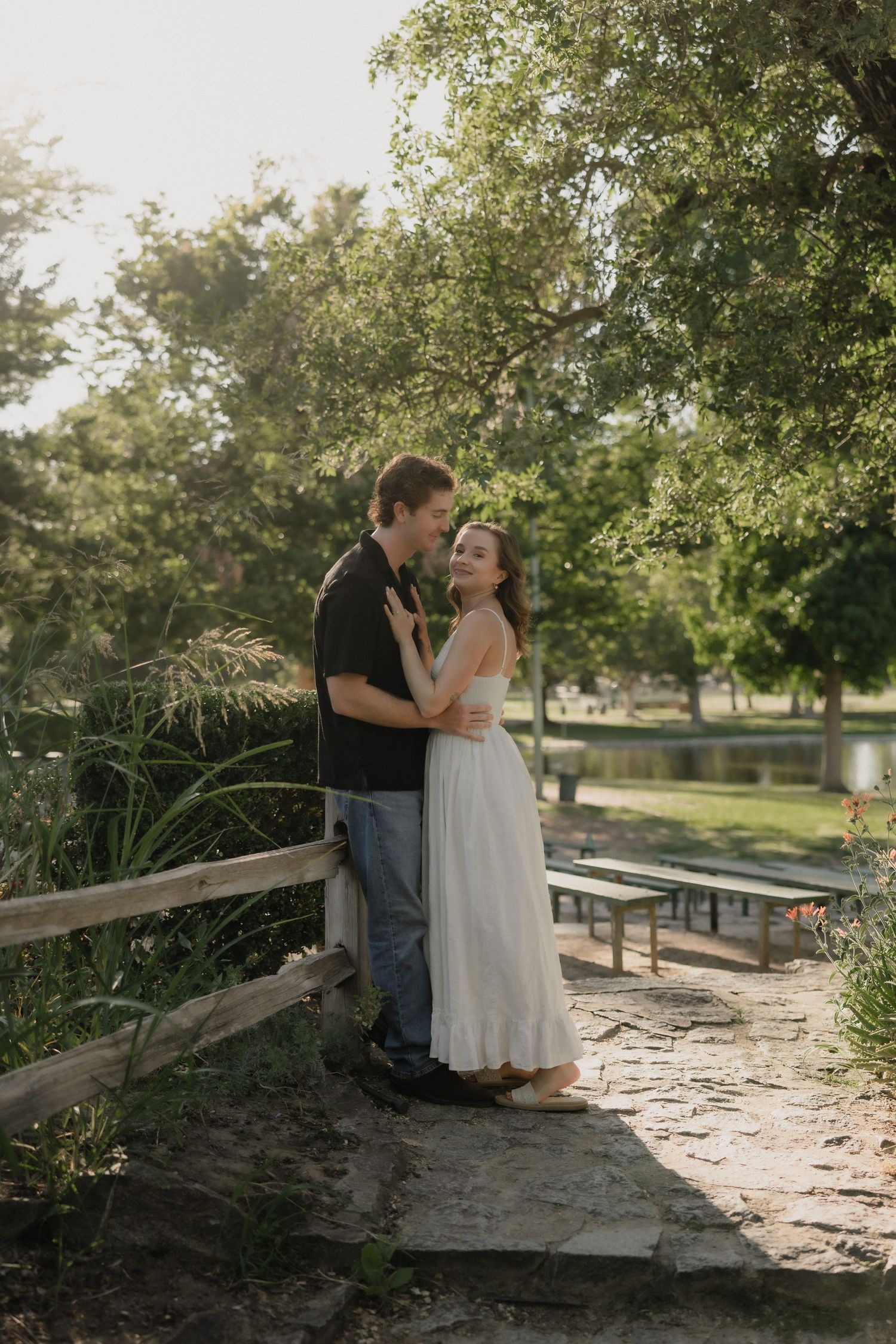 A couple embraces on a stone path in a lush park, the woman in a white dress smiling warmly in golden hour light.