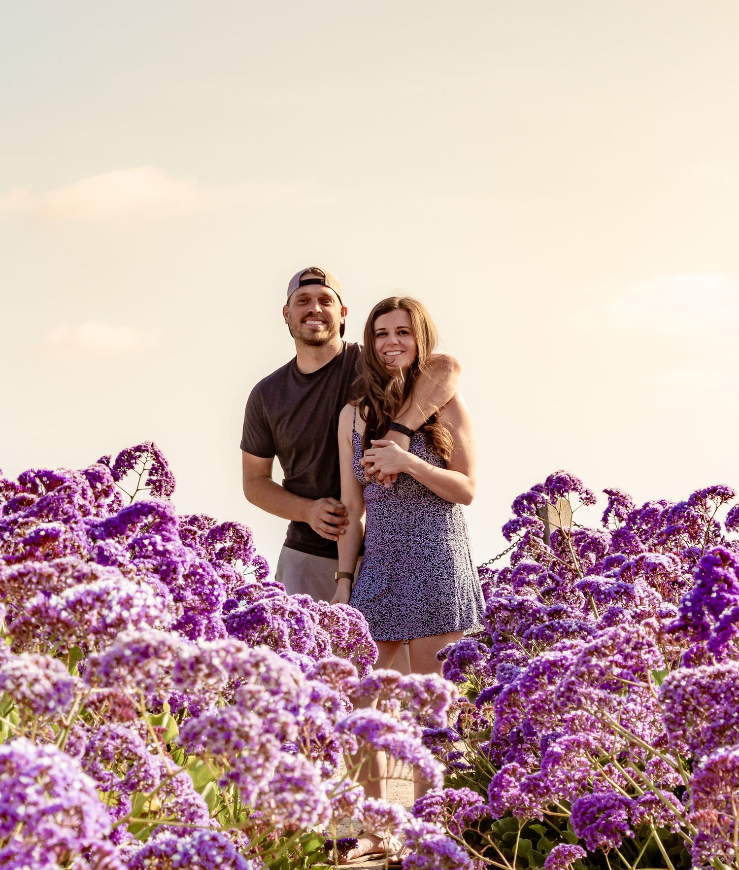 Couple smiling together surrounded by vibrant purple flowers in a sunny outdoor field setting.