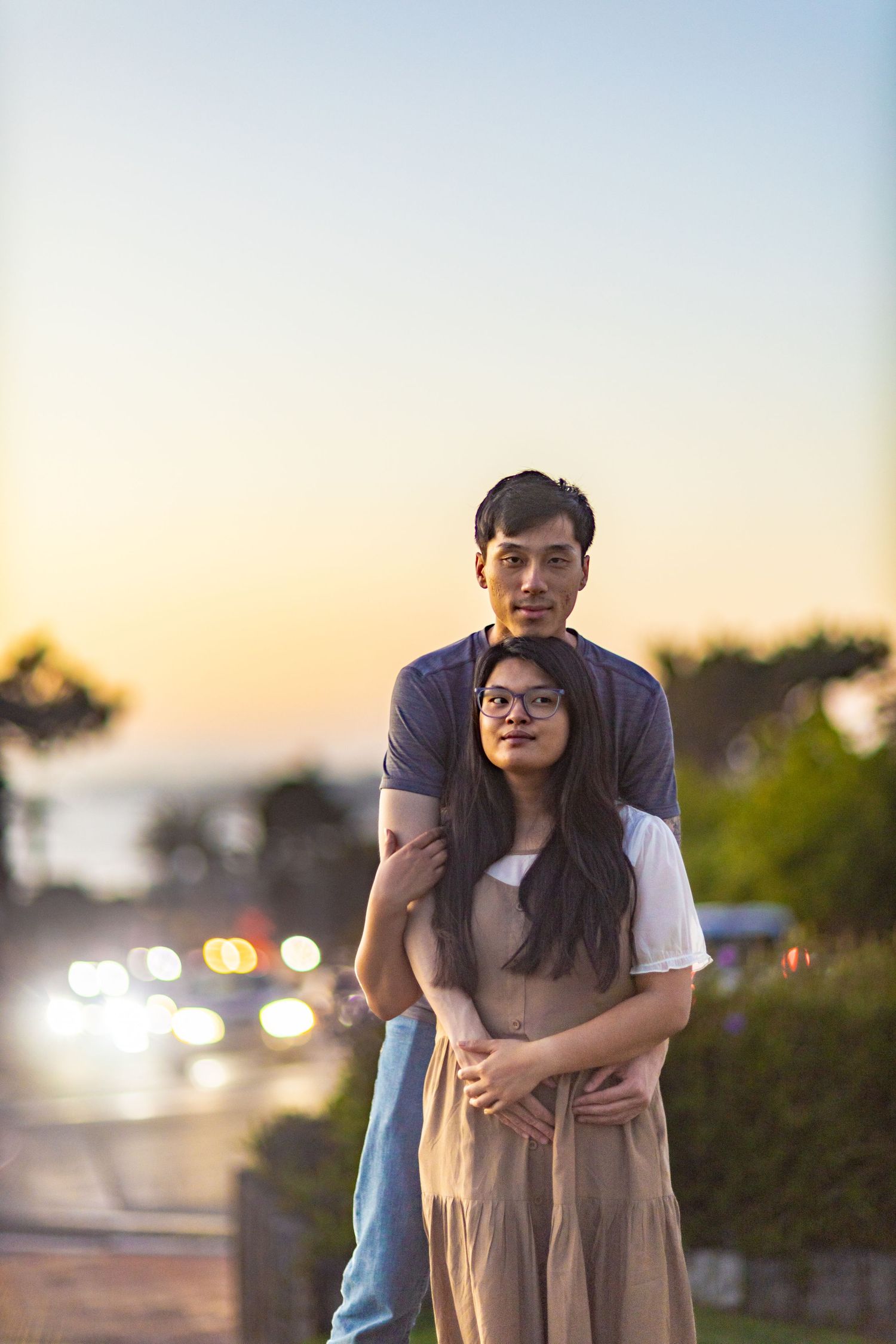 Couple embracing at sunset, man standing behind woman with arms around her, bokeh city lights in background.